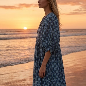 May include: A person wearing a long, flowing blue dress with a floral pattern stands on a beach at sunset. The dress has a V-neck and three-quarter sleeves. The sky is filled with orange and pink hues.