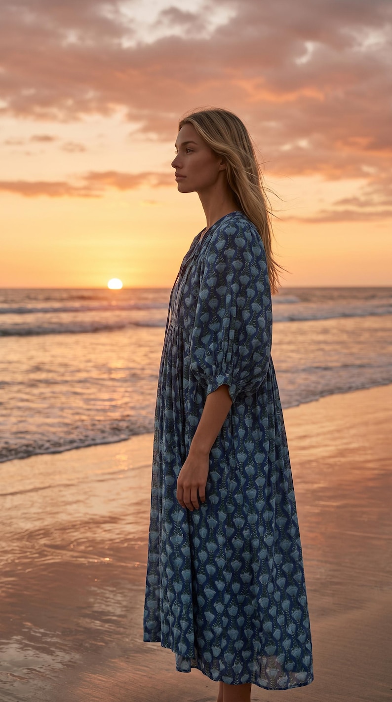 May include: A person wearing a long, flowing blue dress with a floral pattern stands on a beach at sunset. The dress has a V-neck and three-quarter sleeves. The sky is filled with orange and pink hues.