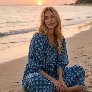 May include: A woman in a blue floral print dress sits cross-legged on a sandy beach at sunset. The dress has a V-neck and long sleeves. The background shows the sea, a setting sun, and palm trees.