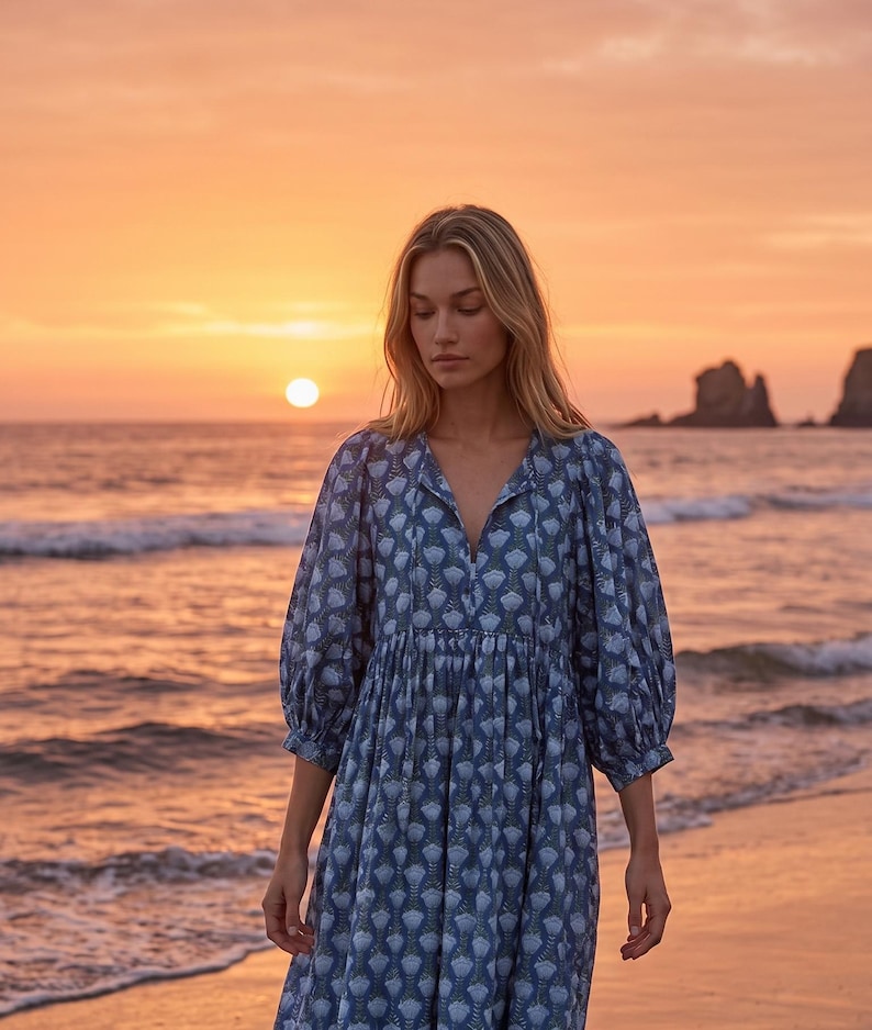 May include: A blue dress with a white floral pattern. The dress features a V-neck, long sleeves, and a gathered waist. The image shows a beach at sunset, with the sun setting over the sea.