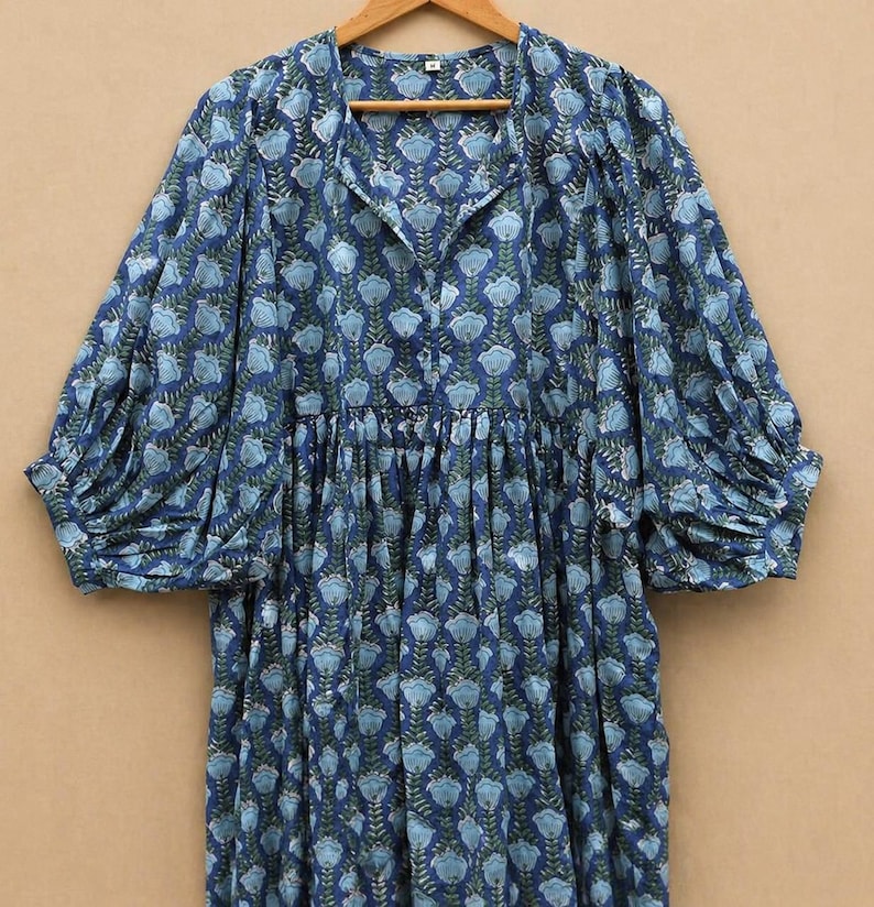 May include: A blue floral print dress with long, voluminous sleeves. The dress has a collared neckline and a gathered waist. The fabric features a pattern of light blue flowers and green leaves on a dark blue background. The dress is hanging on a wooden hanger.