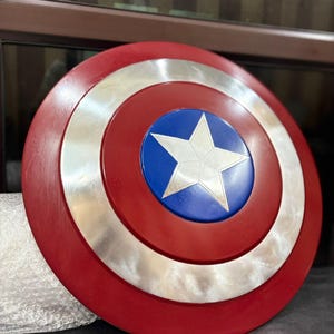 K&ouml;nnte beinhalten: Ein runder Schild mit einem roten Au&szlig;enring, einem silbernen Mittelring und einer blauen Mitte. Ein wei&szlig;er Stern befindet sich in der Mitte des blauen Kreises. Der Schild ist eine Nachbildung von Captain Americas Schild.