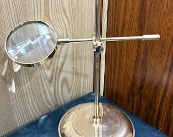 May include: A brass-colored magnifying glass on a stand. The round magnifying glass is held by a horizontal arm, and the stand has a circular base. The stand is approximately 10 inches tall.