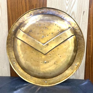 May include: A circular brass shield featuring a prominent V-shaped embossed ridge across the center. The metallic surface displays a polished finish with visible natural oxidation spots, small rivets along the outer rim, and a central point.