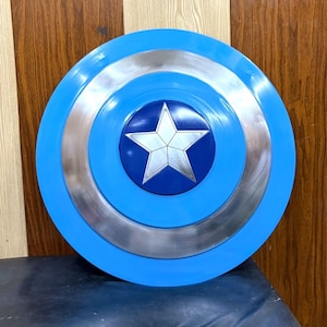 Pode incluir: Um escudo circular com um anel exterior azul brilhante, um anel central prateado metálico e um círculo interior azul escuro. Uma estrela prateada de cinco pontas está centrada no círculo azul escuro, criando um design geométrico distinto.