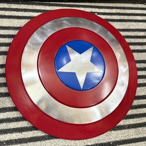 K&ouml;nnte beinhalten: Ein runder Schild mit einem roten Au&szlig;enring, einem silbernen Mittelring und einem blauen Zentrum. Ein wei&szlig;er Stern befindet sich in der Mitte des blauen Kreises. Der Schild ist eine Nachbildung von Captain Americas Schild.
