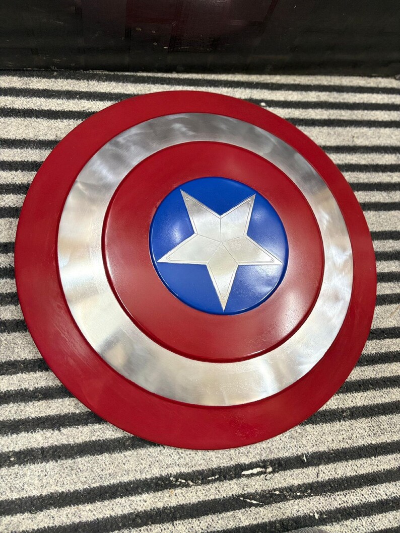 K&ouml;nnte beinhalten: Ein runder Schild mit einem roten Au&szlig;enring, einem silbernen Mittelring und einem blauen Zentrum. Ein wei&szlig;er Stern befindet sich in der Mitte des blauen Kreises. Der Schild ist eine Nachbildung von Captain Americas Schild.