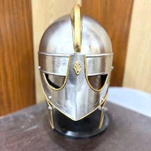 May include: A silver metal helmet with gold detailing. The helmet features a rounded crown, a vertical gold crest, and decorative etching. The face guard has eye openings. The helmet is displayed on a dark surface.
