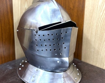 May include: A polished steel knight's helmet with a visor featuring small ventilation holes. The helmet has a rounded top and a neck guard with rivets. The metal reflects the light, creating a bright, metallic appearance. This medieval-style helmet is a decorative item.