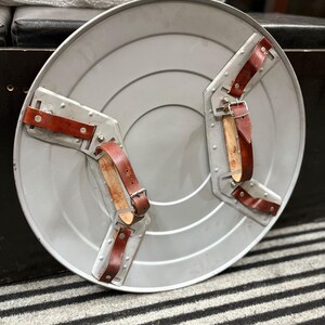 K&ouml;nnte beinhalten: Ein silberfarbener, runder Schild mit einer Reihe konzentrischer Kreise. Er verf&uuml;gt &uuml;ber zwei Metallarmb&auml;nder mit braunen Lederriemen und Schnallen. Der Schild ist in einem klassischen, Vintage-Design gehalten.