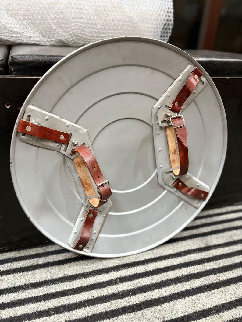 K&ouml;nnte beinhalten: Ein silberfarbener, runder Schild mit einer Reihe konzentrischer Kreise. Er verf&uuml;gt &uuml;ber zwei Metallarmb&auml;nder mit braunen Lederriemen und Schnallen. Der Schild ist in einem klassischen, Vintage-Design gehalten.