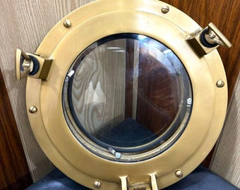 Könnte beinhalten: Rundes Bullauge aus Messing mit einem Fenster aus klarem Glas. Das Bullauge hat mehrere Schrauben und drei Scharnierverschlüsse. Dieser Artikel im nautischen Stil ist für die Montage an einer Wand oder Tür konzipiert.