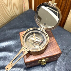 Peut inclure: Une boussole en laiton vintage avec un couvercle à charnière et un miroir intégré. La boussole est posée sur une boîte en bois avec un fermoir en laiton. La boussole a une poignée et un cadran détaillé avec des indications directionnelles.