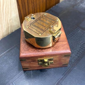 May include: A round brass navigational instrument with a hinged lid featuring an engraved table labelled NATURAL SINE and STANLEY LONDON. The instrument rests on a square wooden box with a brass latch and visible wood grain texture.