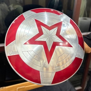 May include: A round, metallic shield with a red and silver design. The shield features a large red star in the center, surrounded by a silver starburst pattern and a red outer rim. The shield is a prop or collectible.