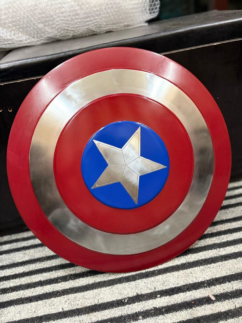 K&ouml;nnte beinhalten: Ein runder Schild mit einem roten Au&szlig;enring, einem silbernen Innenring und einer blauen Mitte. Ein silberner Stern befindet sich in der Mitte des blauen Kreises. Der Schild ist eine Nachbildung des Schildes eines Comic-Superhelden.