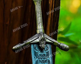 Puede incluir: Espada decorativa con empuñadura ornamentada y hoja azul. La empuñadura presenta intrincados grabados y un acabado bronce. La hoja tiene un diseño azul claro. La espada está apoyada contra un tronco de árbol. El texto "ARVAK STEEL" es visible.