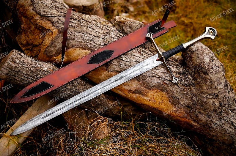 May include: A detailed image of a sword with a patterned blade and a black handle. The sword rests on a wooden log alongside a brown leather sheath with black accents. The sword's hilt has a silver finish with gold details.