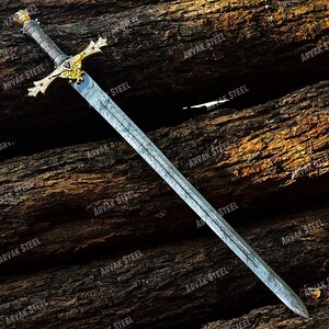 May include: A long, ornate sword with a patterned blade and a gold-colored hilt. The hilt features intricate designs and red accents. The sword is displayed against a backdrop of textured, dark brown wood. The text "ARVAK STEEL" is visible on the blade.