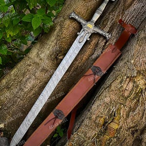 May include: A detailed image of a medieval sword with a patterned blade and ornate silver hilt. The sword rests beside a brown leather sheath, secured with a leather strap. The background is a tree trunk and green foliage.