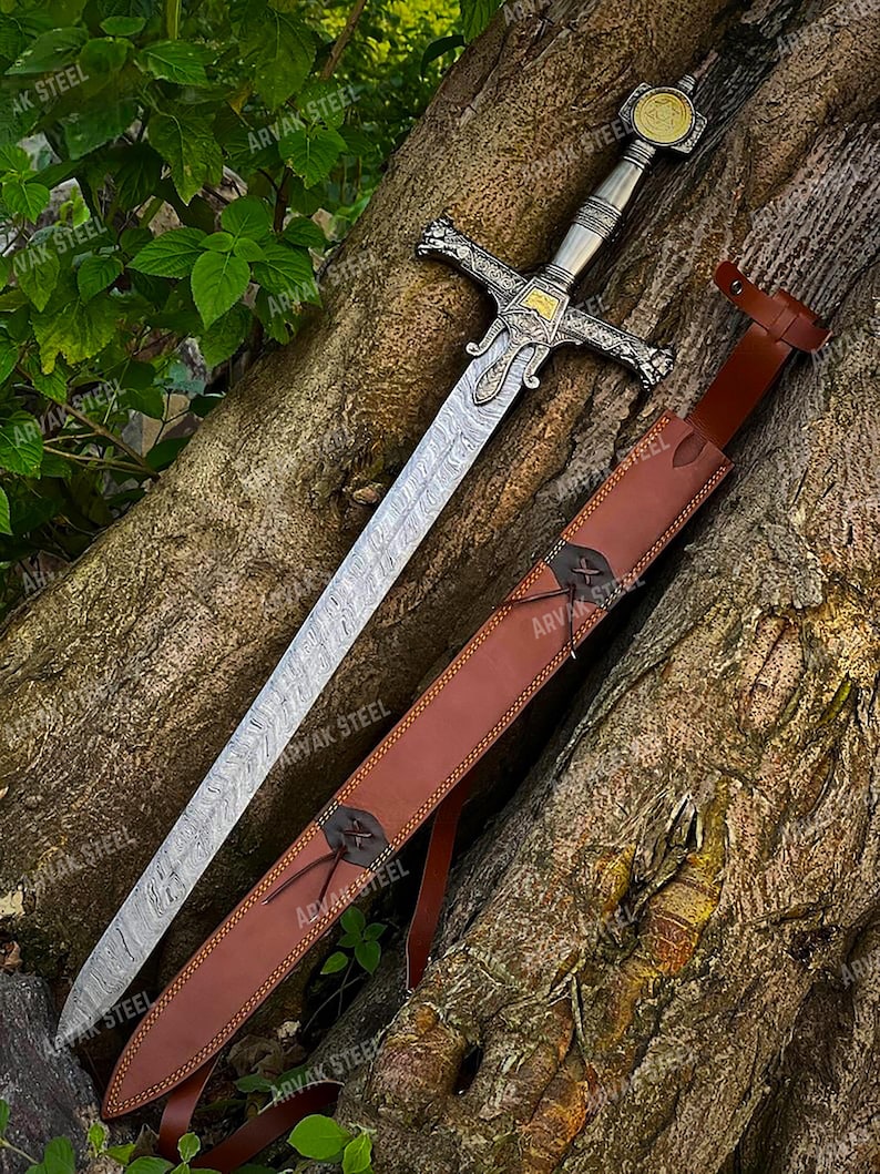 May include: A detailed image of a medieval sword with a patterned blade and ornate silver hilt. The sword rests beside a brown leather sheath, secured with a leather strap. The background is a tree trunk and green foliage.