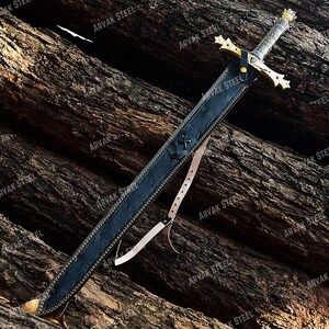 May include: A medieval-style sword with a gold crossguard and a silver handle, resting in a black leather sheath. The sheath has a decorative stitch pattern and a leather strap. The sword is displayed on a textured wooden surface.