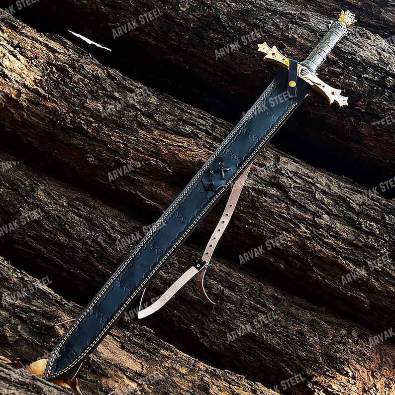 May include: A medieval-style sword with a gold crossguard and a silver handle, resting in a black leather sheath. The sheath has a decorative stitch pattern and a leather strap. The sword is displayed on a textured wooden surface.