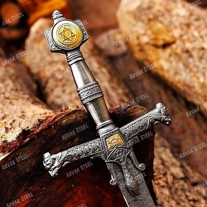 May include: A detailed close-up of a decorative sword with a silver handle and a gold-colored emblem featuring a Star of David. The blade has a patterned design. The sword rests on a wooden surface, with the text "ARVAK STEEL" visible.