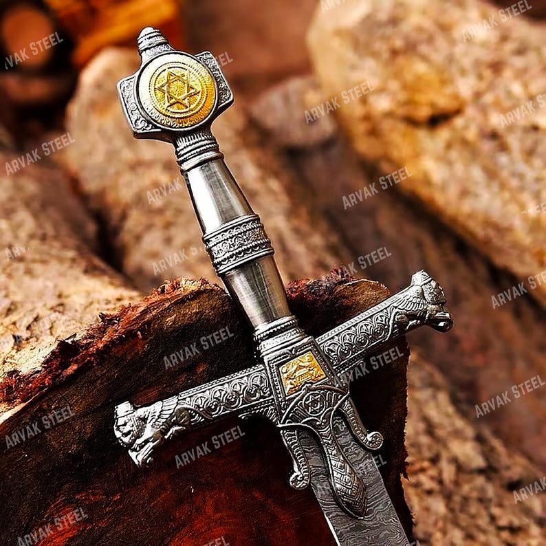 May include: A detailed close-up of a decorative sword with a silver handle and a gold-colored emblem featuring a Star of David. The blade has a patterned design. The sword rests on a wooden surface, with the text "ARVAK STEEL" visible.