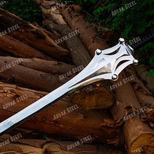 May include: A polished steel sword with a decorative hilt and a black wrapped handle. The blade is long and sharp, resting on a pile of wooden logs. The text "ARVAK STEEL" is visible on the image.