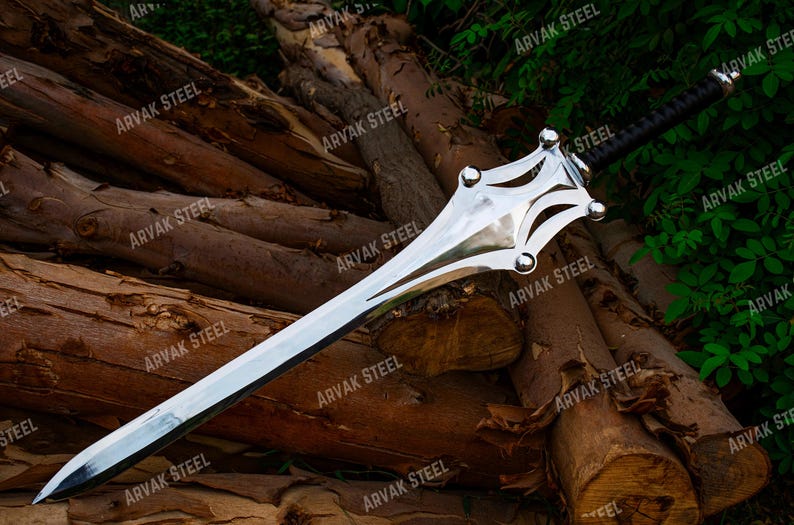 May include: A polished steel sword with a decorative hilt and a black wrapped handle. The blade is long and sharp, resting on a pile of wooden logs. The text "ARVAK STEEL" is visible on the image.