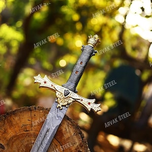 May include: A decorative sword with a gold and red hilt, a black handle, and a patterned blade. The sword is resting on a wooden log, with a blurred background of green foliage and sunlight.