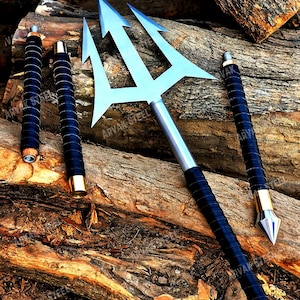 May include: A silver trident with three prongs and a black wrapped handle, along with two additional handles. The text "ARVAK STEEL" is visible on the handles. The items are displayed on a wooden surface.