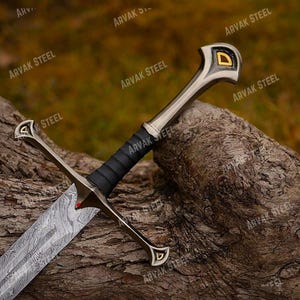 May include: A detailed image of a sword with a patterned blade and a black handle. The sword has a silver hilt with gold accents and rests on a textured, brown surface. The sword's design suggests a historical or fantasy theme.