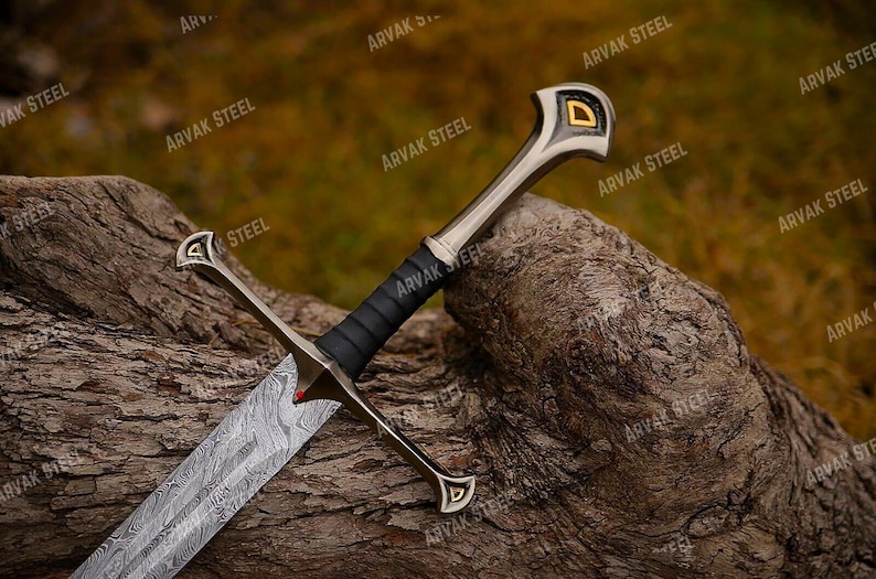 May include: A detailed image of a sword with a patterned blade and a black handle. The sword has a silver hilt with gold accents and rests on a textured, brown surface. The sword's design suggests a historical or fantasy theme.