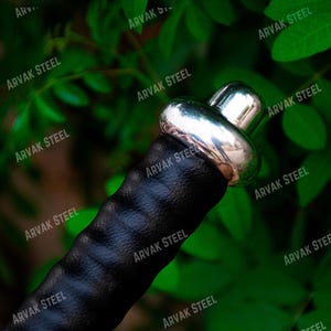 May include: Close-up of a sword hilt with a black leather grip and a shiny silver metal pommel. The hilt is set against a backdrop of green foliage, creating a contrast of textures and colors. The text "ARVAK STEEL" is visible.