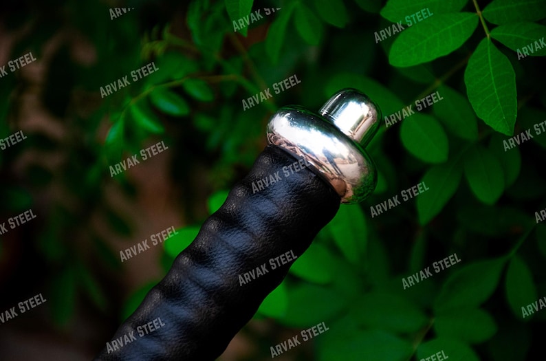 May include: Close-up of a sword hilt with a black leather grip and a shiny silver metal pommel. The hilt is set against a backdrop of green foliage, creating a contrast of textures and colors. The text "ARVAK STEEL" is visible.