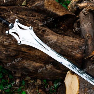 May include: A silver-colored sword with a black handle and a unique, decorative hilt. The blade is long and sharp, with a polished finish. The sword is displayed on a background of weathered wood.