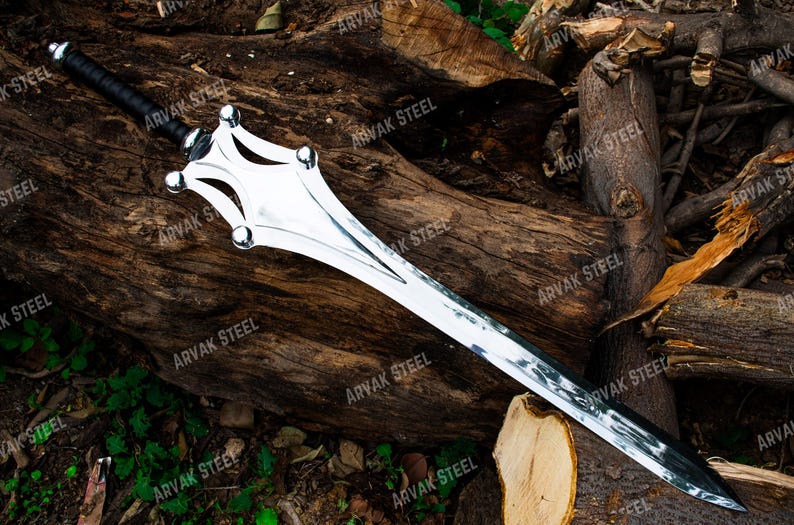 May include: A silver-colored sword with a black handle and a unique, decorative hilt. The blade is long and sharp, with a polished finish. The sword is displayed on a background of weathered wood.