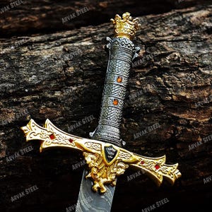 May include: A decorative sword with a gold-colored hilt and a silver-colored handle. The hilt features a detailed design with red accents and a crest. The handle is adorned with orange-colored gems and a crown at the top. The blade has a patterned design.