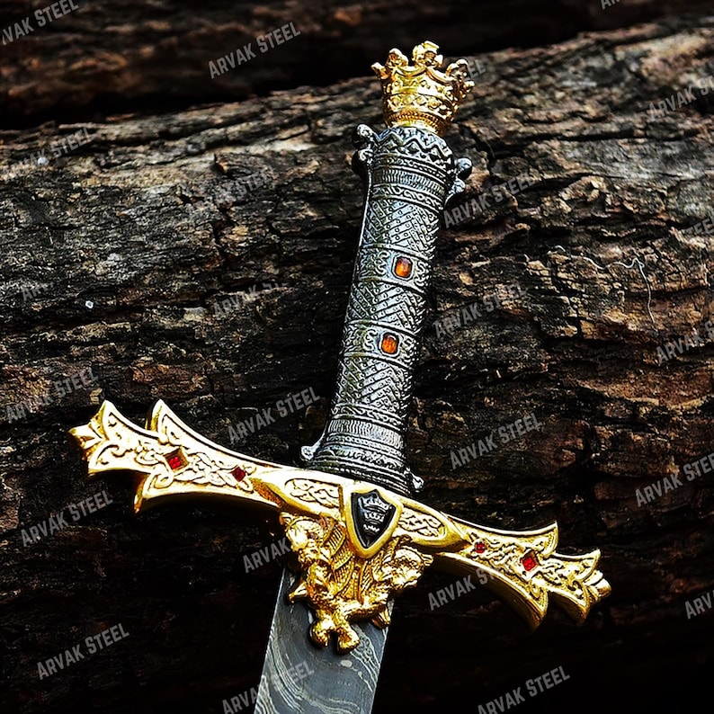 May include: A decorative sword with a gold-colored hilt and a silver-colored handle. The hilt features a detailed design with red accents and a crest. The handle is adorned with orange-colored gems and a crown at the top. The blade has a patterned design.