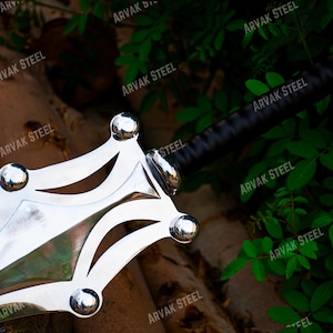 May include: A close-up of a polished steel sword with a black handle. The blade has a unique, decorative design with rounded accents. The sword is set against a backdrop of wood and green foliage.