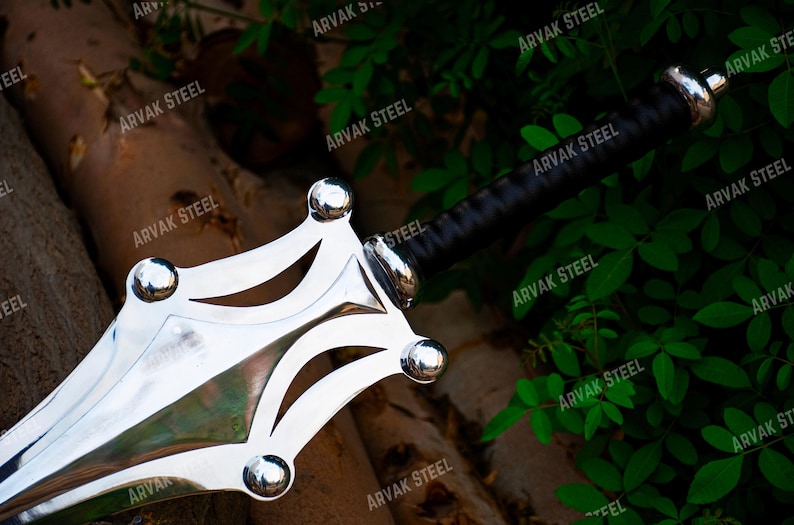 May include: A close-up of a polished steel sword with a black handle. The blade has a unique, decorative design with rounded accents. The sword is set against a backdrop of wood and green foliage.