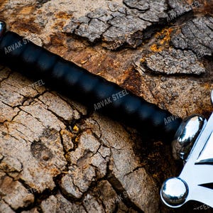May include: A close-up of a sword with a black handle and a silver blade. The handle is wrapped in black leather. The blade has a decorative design. The sword is resting on a textured, brown surface.