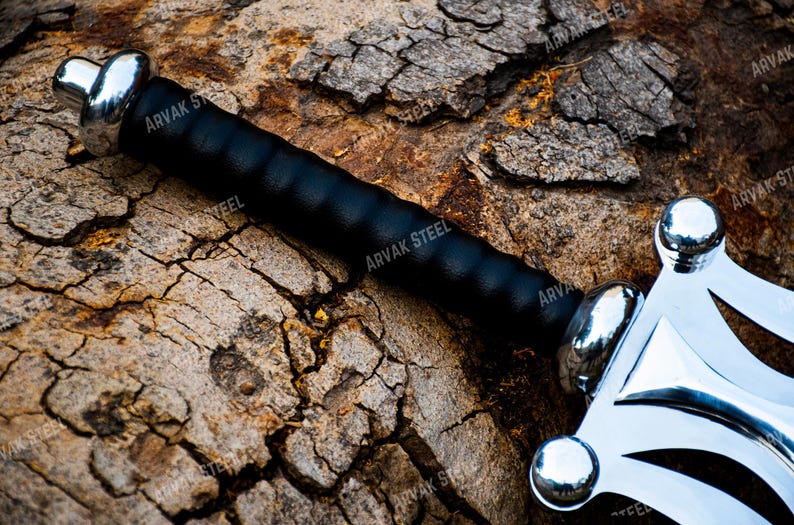 May include: A close-up of a sword with a black handle and a silver blade. The handle is wrapped in black leather. The blade has a decorative design. The sword is resting on a textured, brown surface.