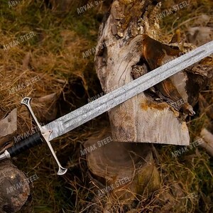 May include: A detailed image of a long sword with a silver blade and a black and silver handle. The sword is resting on a wooden surface with a background of dried leaves and wood.