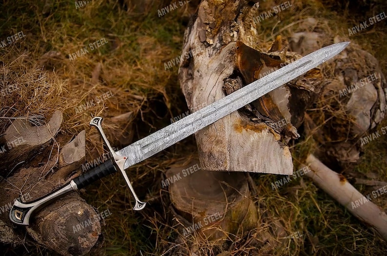 May include: A detailed image of a long sword with a silver blade and a black and silver handle. The sword is resting on a wooden surface with a background of dried leaves and wood.