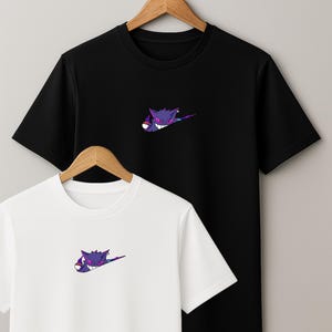 Peut inclure: Deux t-shirts à manches courtes, un noir et un blanc, chacun avec un motif de fantôme de dessin animé violet et bleu sur la poitrine. Les chemises sont suspendues à un cintre en bois sur un fond neutre.