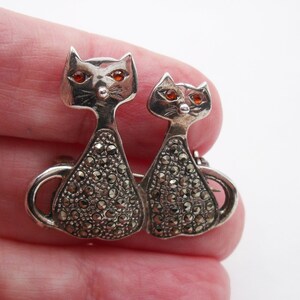 May include: A silver-toned cat brooch featuring two stylized cat figures. The cats have orange gemstone eyes and bodies encrusted with small, sparkling stones. The brooch is a vintage piece, likely made of sterling silver.