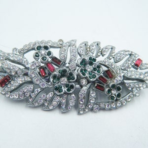 May include: A silver floral brooch with clear, red, and green gemstones. The brooch has a symmetrical design of leaves and flowers, with the gemstones adding sparkle and colour. A vintage-inspired accessory.